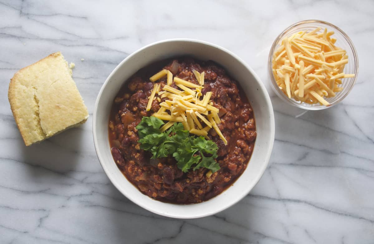 Vegan Tofu Chili in a bowl with a side of shredded vegan cheddar cheese and vegan cornbread
