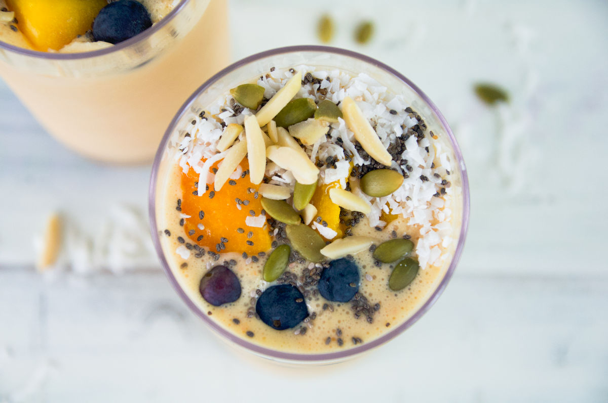 Close up of mango smoothie in a tall glass topped with blueberries, mango cubes, pumpkin seeds, almonds slices, coconut flakes, and chia seeds on a white background