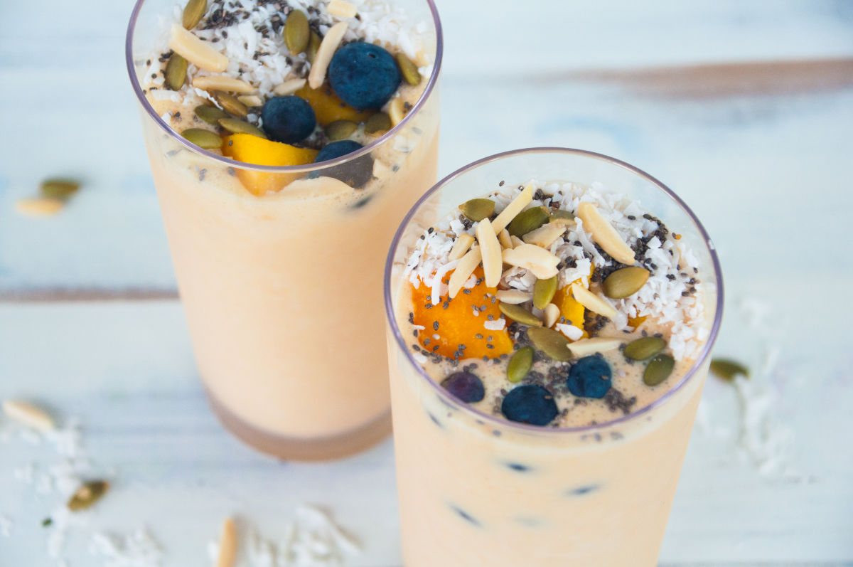 Two mango smoothies in tall glasses topped with blueberries, mango cubes, pumpkin seeds, almonds slices, coconut flakes, and chia seeds on a white background