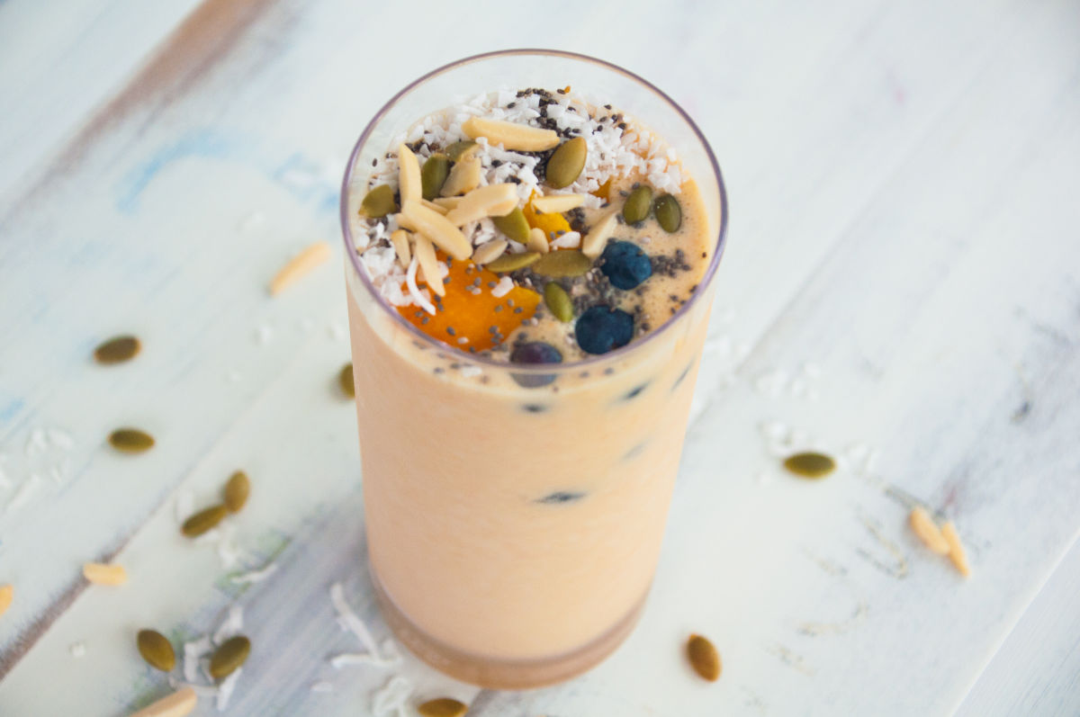Mango smoothie in a tall glass topped with blueberries, mango cubes, pumpkin seeds, almonds slices, coconut flakes, and chia seeds on a white background