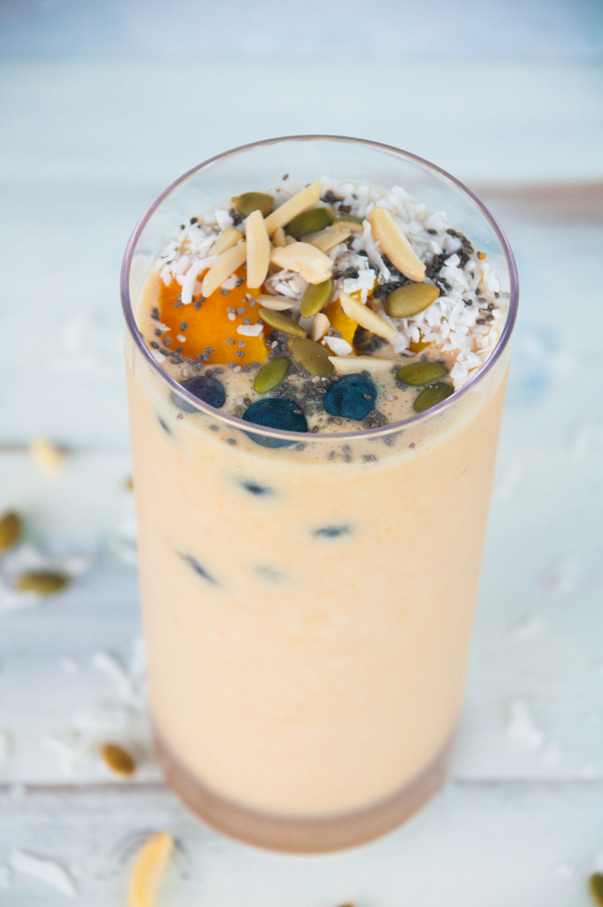 Mango smoothie in a tall glass topped with blueberries, mango cubes, pumpkin seeds, almonds slices, coconut flakes, and chia seeds on a white background
